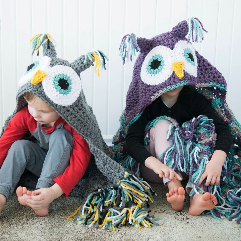 May include: Two children wearing crocheted owl hooded blankets. The blankets are gray and purple with colorful fringe. The children are sitting on a white surface.