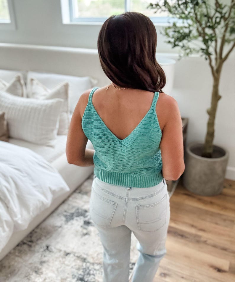 Pu&ograve; includere: Un top all'uncinetto turchese con scollo a V. Il top ha spalline sottili e un orlo a coste. La persona indossa jeans di denim lavati chiari. Lo sfondo include un letto con cuscini bianchi e una pianta in vaso.