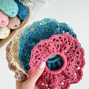 Può includere: Una collezione di scrunchies all'uncinetto fatti a mano in vari colori, tra cui rosa, blu e beige. Gli scrunchies hanno un bordo smerlato e sono realizzati con filato morbido. Sullo sfondo, una ciotola di legno con filato dai colori coordinati.