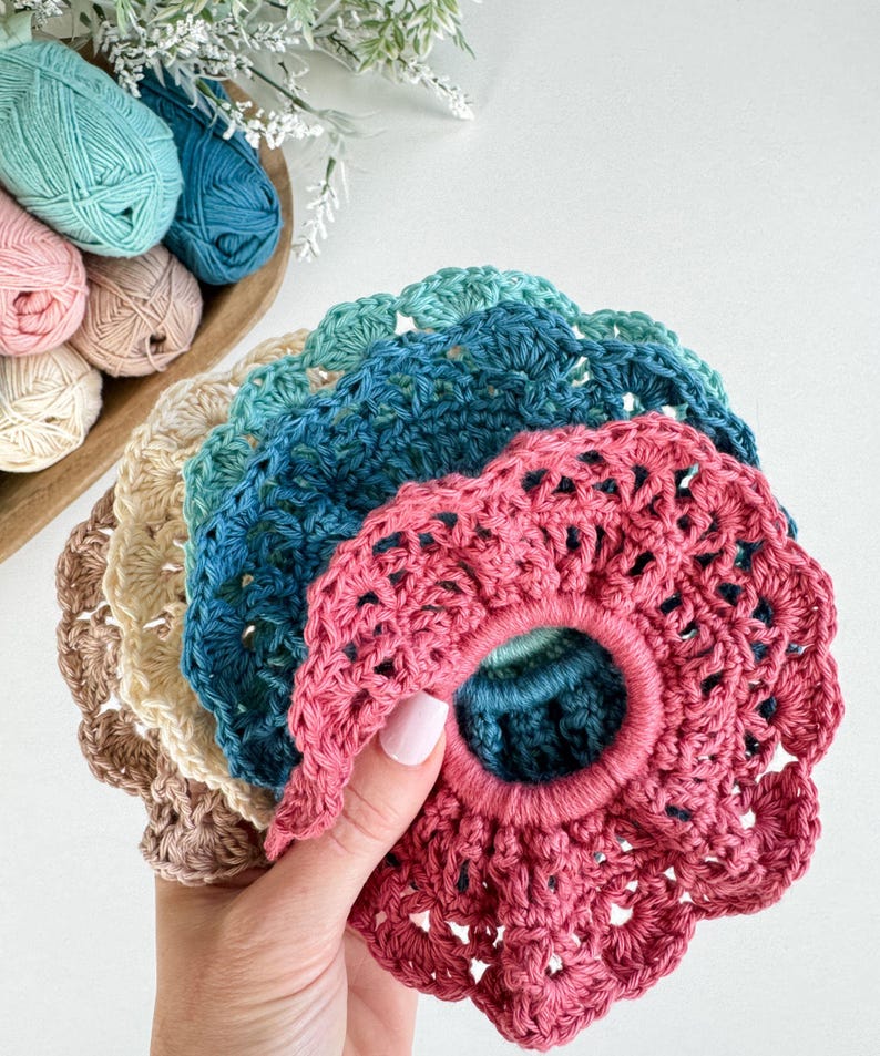 May include: A collection of handmade crochet scrunchies in various colors, including pink, blue, and beige. The scrunchies have a scalloped edge design and are made with soft yarn. A wooden bowl of yarn in coordinating colors is in the background.