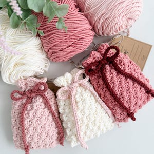 May include: A selection of handmade crochet items in pink and white. The image shows several balls of yarn and three small drawstring bags with matching cord ties. The bags are in varying shades of pink and white.