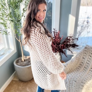 May include: A cream-colored, open-weave crochet cardigan with long sleeves. The cardigan has a relaxed fit and a textured appearance. The garment is knee-length and appears to be made of a soft, natural fiber. The style is casual and suitable for layering.