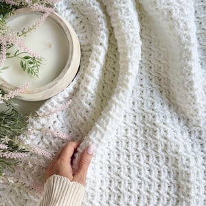 Könnte beinhalten: Eine weiße, strukturierte Häkeldecke mit Zopfmuster. Die Decke ist gefaltet und zeigt das komplizierte Stichmuster. Eine Holzschale mit einer weißen Kerze und Zweigen mit Grün und rosa Blüten befindet sich im Hintergrund.