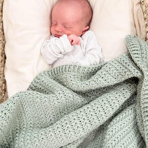 Puede incluir: Un reci&eacute;n nacido con los ojos cerrados descansa sobre una almohada de color crema, vistiendo una camisa blanca de manga larga acanalada. Una manta de ganchillo verde salvia con un patr&oacute;n de estrellas texturizado cubre el cuerpo.
