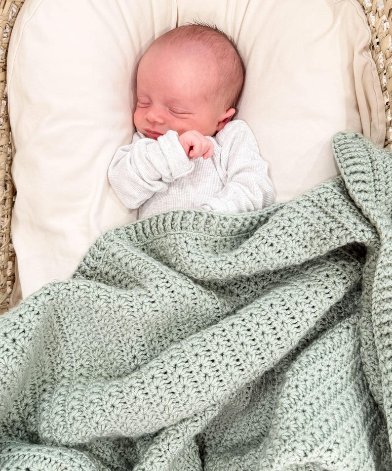Puede incluir: Un reci&eacute;n nacido con los ojos cerrados descansa sobre una almohada de color crema, vistiendo una camisa blanca de manga larga acanalada. Una manta de ganchillo verde salvia con un patr&oacute;n de estrellas texturizado cubre el cuerpo.