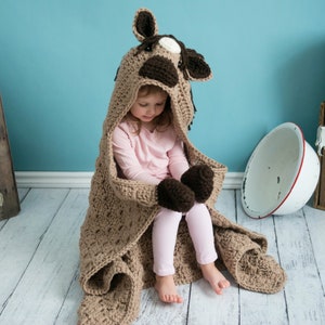 Pu&ograve; includere: Una coperta di cavallo in crochet marrone con cappuccio e guanti. La coperta &egrave; realizzata con un punto testurizzato e ha un viso di cavallo marrone e bianco sul cappuccio.