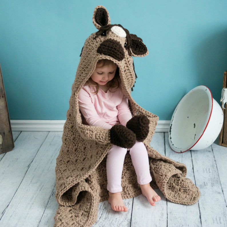 Pu&ograve; includere: Una coperta di cavallo in crochet marrone con cappuccio e guanti. La coperta &egrave; realizzata con un punto testurizzato e ha un viso di cavallo marrone e bianco sul cappuccio.