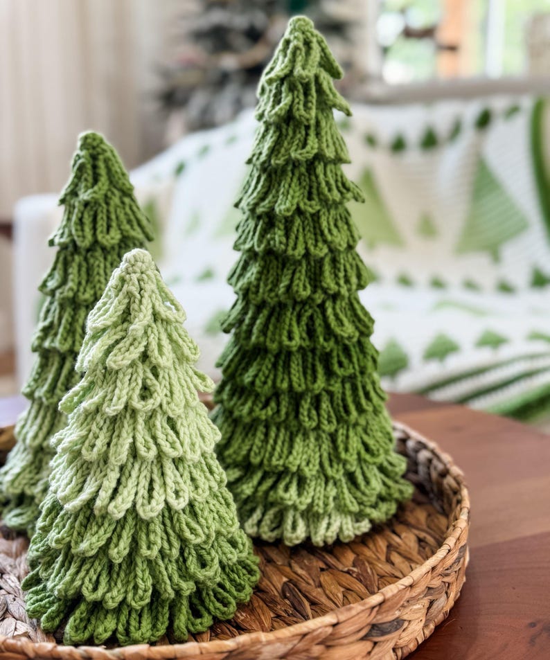 May include: Three handmade green knitted Christmas trees of varying sizes sit in a woven basket. The trees have a textured, layered design, with a lighter green tree in the front and darker green trees behind it. The background includes a white blanket with green tree designs.