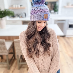 May include: A purple, blue, and gray crocheted beanie with a fluffy pom pom on top. The beanie has a brown leather patch with the text "A&M" on it.
