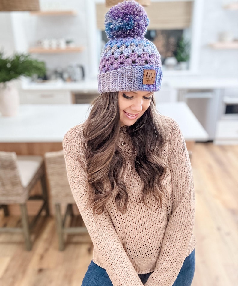 May include: A purple, blue, and gray crocheted beanie with a fluffy pom pom on top. The beanie has a brown leather patch with the text "A&M" on it.