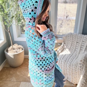 May include: A light blue crochet cardigan with a hood and a pocket. The cardigan is made with a variegated yarn that has shades of blue, pink, and purple.