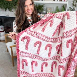 K&ouml;nnte beinhalten: Eine wei&szlig;-rote geh&auml;kelte Decke mit einem sich wiederholenden Muster aus Zuckerstangen. Die Decke hat einen roten Rand und wird von einer Person gehalten. Ein festlicher Artikel.