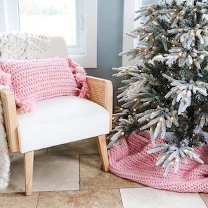 Puede incluir: Un sillón blanco con un cojín de punto rosa y flecos. El sillón está delante de una ventana y un árbol de Navidad nevado con luces blancas.