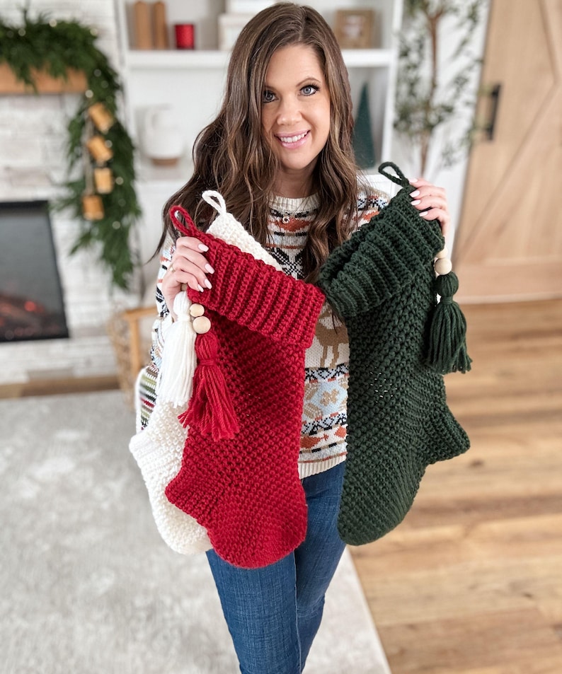 May include: Three handmade Christmas stockings in red, white, and green. Each stocking has a knitted texture, a tassel, and wooden bead accents. The red stocking is in the center, with the white and green stockings on either side.