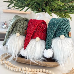 May include: Three crocheted gnomes with white fur beards, wearing green, red, and teal hats. The gnomes are sitting in a wooden bowl on a white surface.