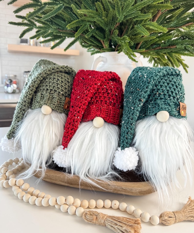 May include: Three crocheted gnomes with white fur beards, wearing green, red, and teal hats. The gnomes are sitting in a wooden bowl on a white surface.