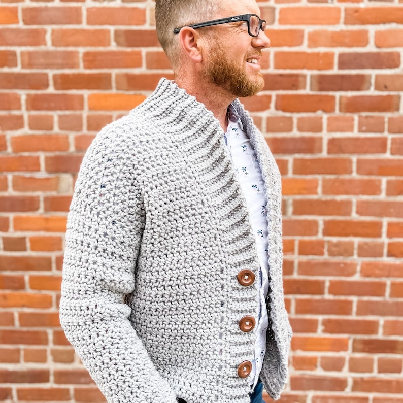 May include: A grey crocheted cardigan with a button fastening and a collar. The cardigan is being worn by a person with a beard and glasses.