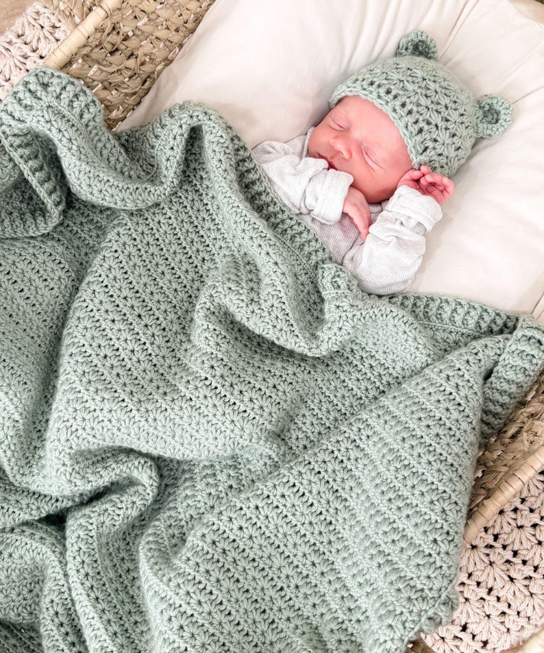 Puede incluir: Una manta de beb&eacute; tejida a ganchillo en color verde salvia con un patr&oacute;n de punto de estrella texturizado, junto con un gorro de beb&eacute; a juego en verde salvia con peque&ntilde;as orejas redondeadas. El conjunto est&aacute; hecho de hilo y es uniforme.