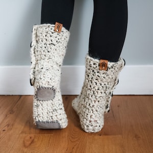 May include: A pair of white crocheted slipper boots with brown leather accents and a grey suede sole. The boots have a button detail on the side.