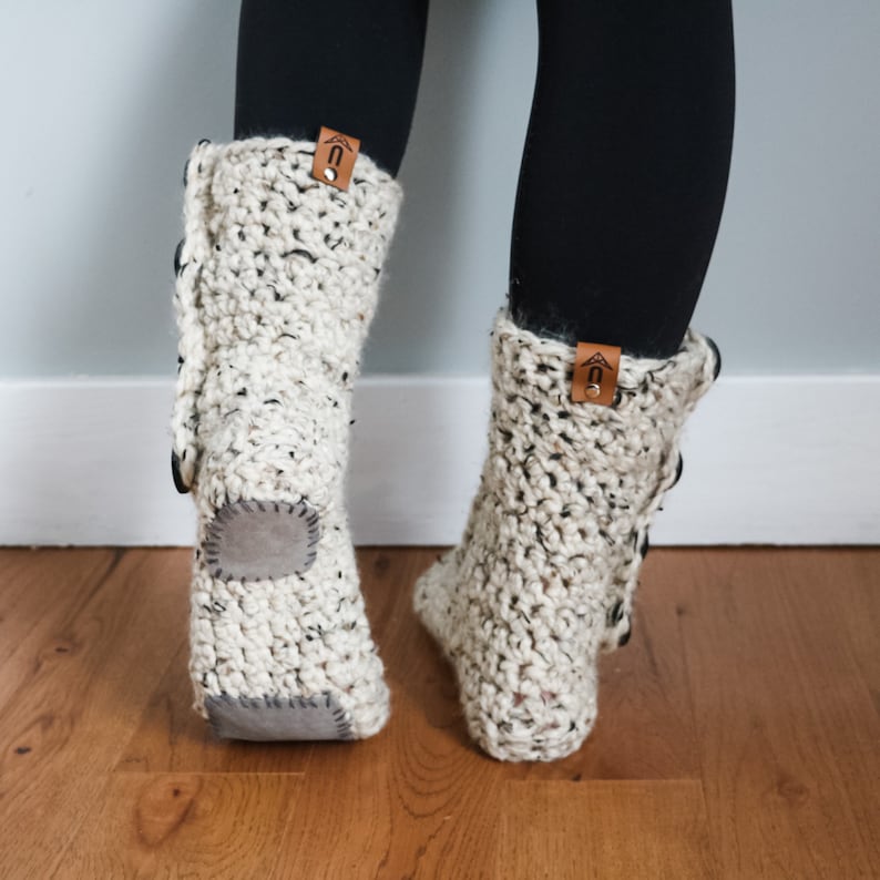 May include: A pair of white crocheted slipper boots with brown leather accents and a grey suede sole. The boots have a button detail on the side.