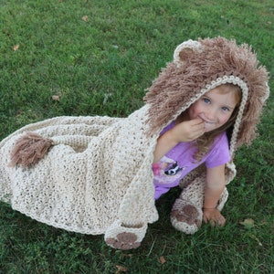 May include: A child wearing a crocheted lion hooded blanket with a brown and beige color scheme. The blanket has a tail and paw prints.