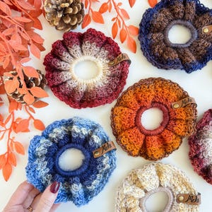 May include: A collection of handmade crochet scrunchies in various colors, including red, blue, orange, and cream. Each scrunchie features a unique color gradient and is adorned with a small leather tag that reads "Mds". The scrunchies are arranged on a white surface with fall-themed decorations.