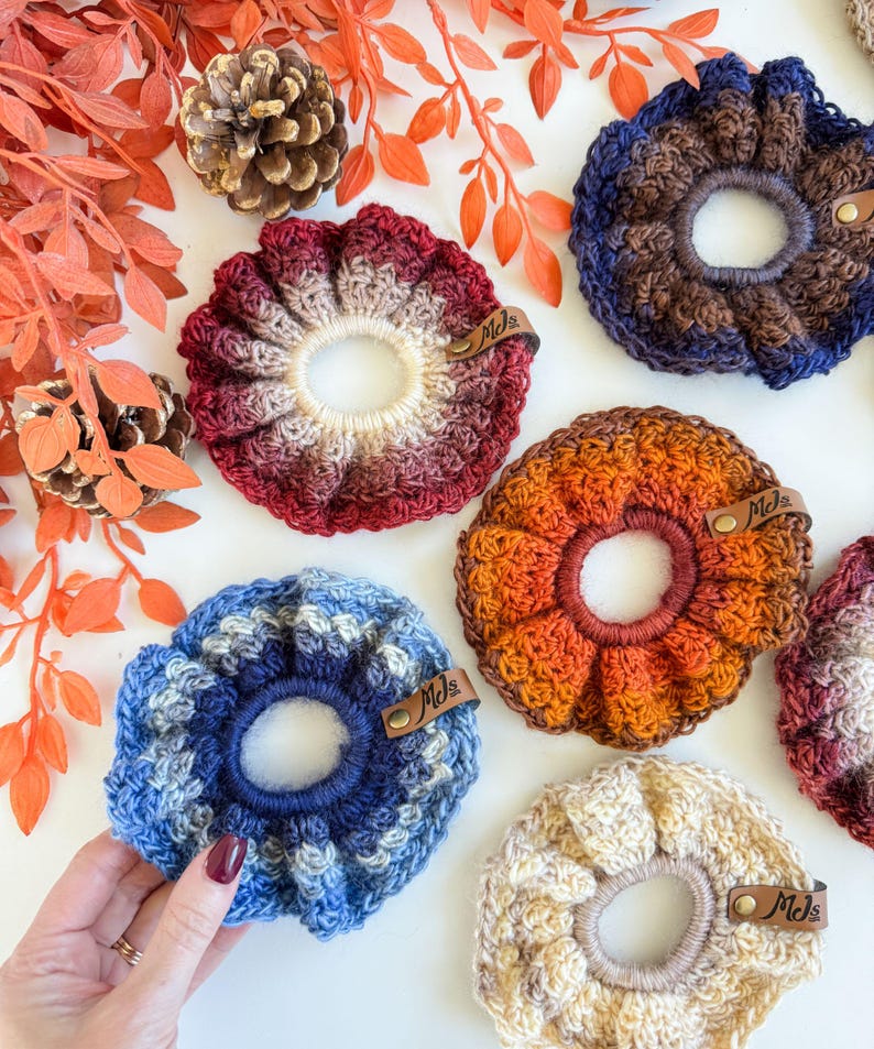 May include: A collection of handmade crochet scrunchies in various colors, including red, blue, orange, and cream. Each scrunchie features a unique color gradient and is adorned with a small leather tag that reads "Mds". The scrunchies are arranged on a white surface with fall-themed decorations.