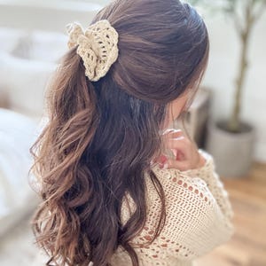 May include: A beige crochet hair clip is worn in the hair. The hair clip is made of a woven, open-work design. The hair is long, wavy, and brown. The person is wearing a beige crochet sweater.