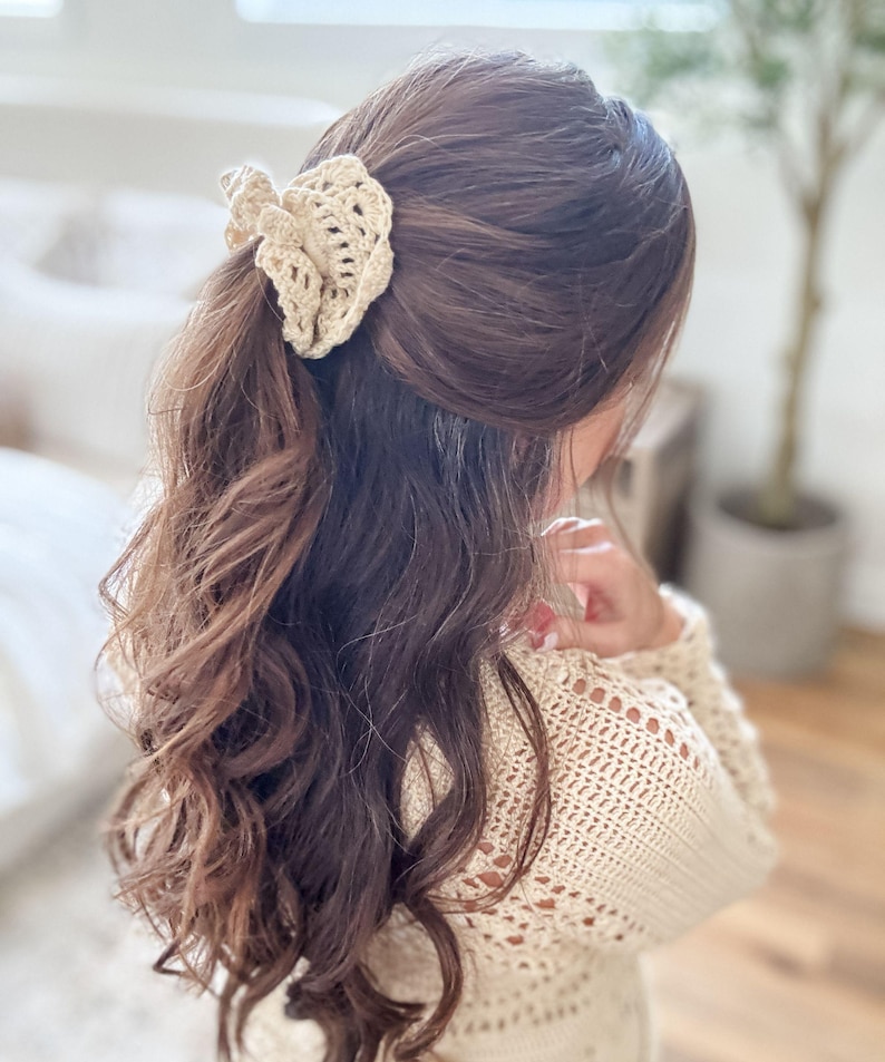 May include: A beige crochet hair clip is worn in the hair. The hair clip is made of a woven, open-work design. The hair is long, wavy, and brown. The person is wearing a beige crochet sweater.