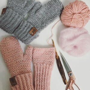May include: Two pairs of crocheted mittens, one pair in gray and one pair in pink. The mittens are laid out on a white surface with yarn and a pair of scissors.