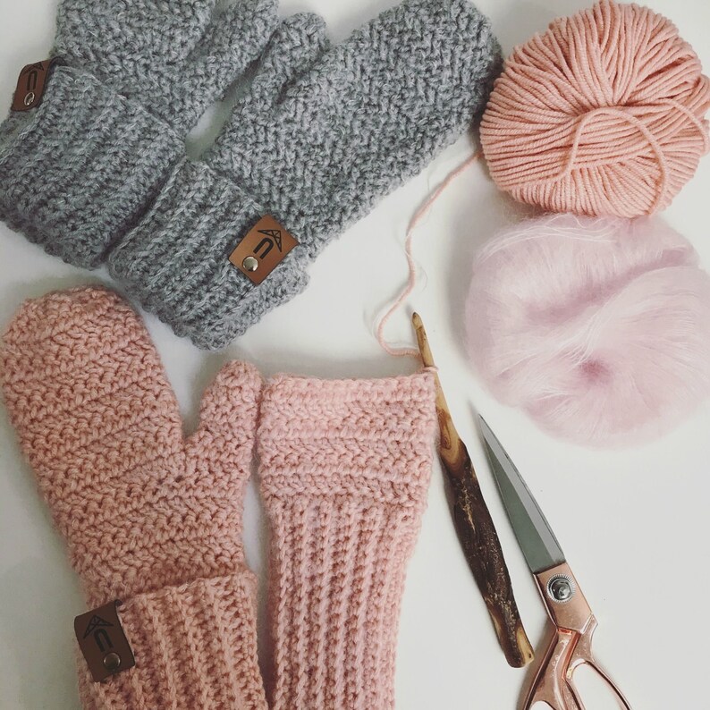 May include: Two pairs of crocheted mittens, one pair in gray and one pair in pink. The mittens are laid out on a white surface with yarn and a pair of scissors.