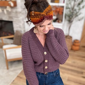 May include: A purple crochet cardigan with gold buttons is paired with blue jeans. A colorful, striped crochet headband in shades of orange, brown, and red is worn on the head. The headband has a small leather tag.