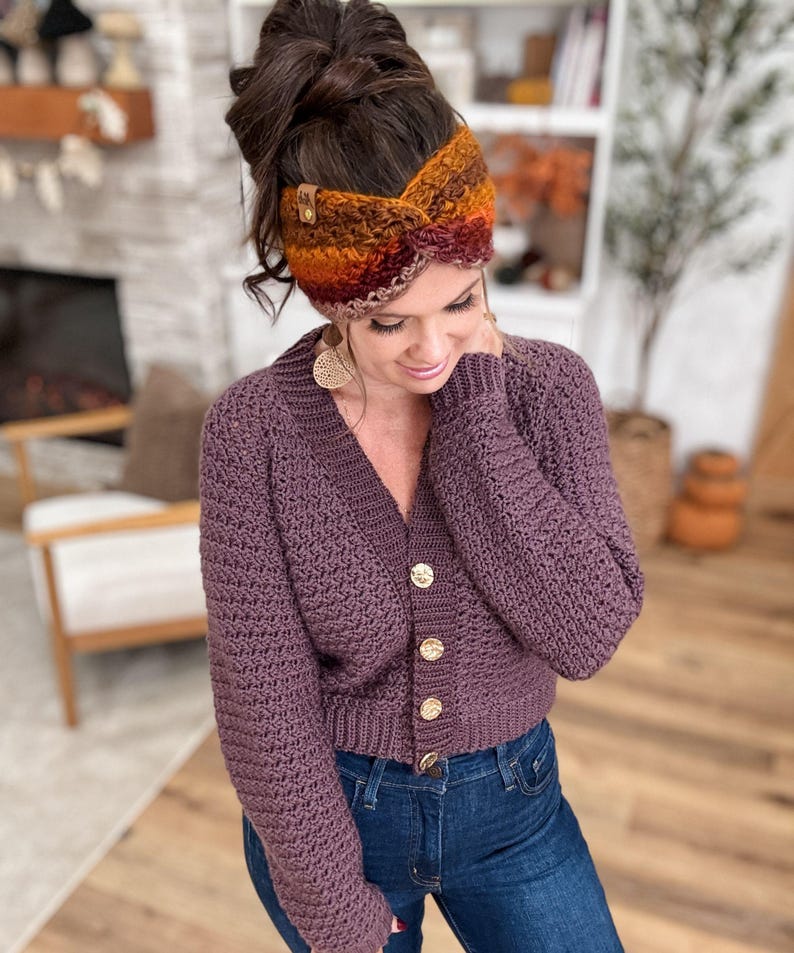 May include: A purple crochet cardigan with gold buttons is paired with blue jeans. A colorful, striped crochet headband in shades of orange, brown, and red is worn on the head. The headband has a small leather tag.