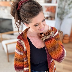 May include: A hooded, crochet cardigan in shades of orange, red, and cream. The long-sleeved sweater has a striped pattern and is paired with a matching scrunchie and bracelet. The cardigan is open in the front and has a relaxed fit.