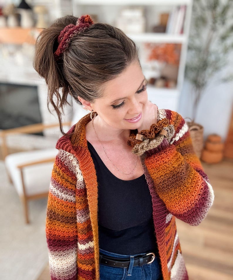 May include: A hooded, crochet cardigan in shades of orange, red, and cream. The long-sleeved sweater has a striped pattern and is paired with a matching scrunchie and bracelet. The cardigan is open in the front and has a relaxed fit.