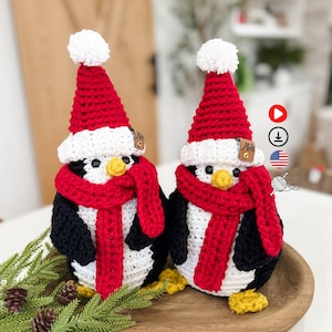 Puede incluir: Dos adornos de pingüino hechos a mano con ganchillo. Cada pingüino tiene un cuerpo negro y blanco, una bufanda y un gorro rojos con un pompón blanco y pies amarillos. Los pingüinos se exhiben en una bandeja de madera con agujas de pino y piñas.