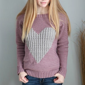 Puede incluir: Un su&eacute;ter de crochet malva con un dise&ntilde;o de coraz&oacute;n gris. El su&eacute;ter tiene mangas largas y un cuello redondo.