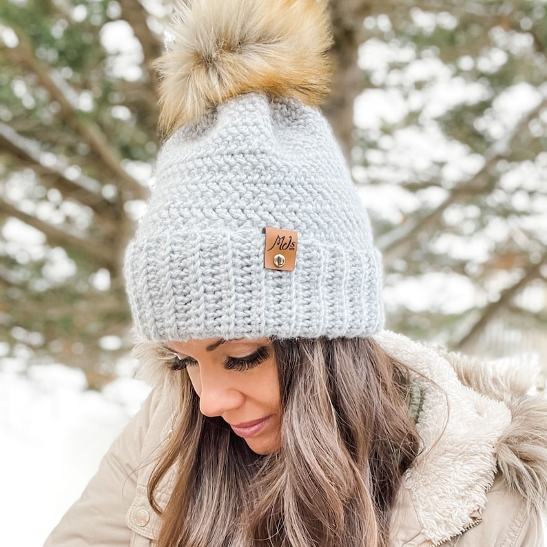 May include: A gray crocheted beanie with a faux fur pom pom and a brown leather label that says "Mels".