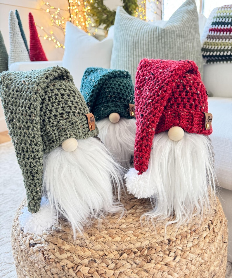 May include: Three crocheted gnomes with white beards and different colored hats. The gnome on the left has a green hat, the gnome in the middle has a red hat, and the gnome on the right has a green hat. The gnomes are sitting on a woven basket.
