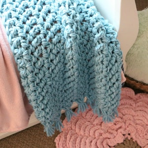 Pu&ograve; includere: Una coperta all'uncinetto blu chiaro con un bordo a frangia. La coperta &egrave; drappeggiata su una sedia bianca e un tappeto all'uncinetto rosa.