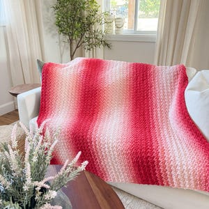 May include: A handmade, textured blanket in shades of red and pink, draped over a white sofa. The blanket features a gradient effect, transitioning from light pink to a vibrant red. The cozy throw adds a touch of warmth and color to the living space.