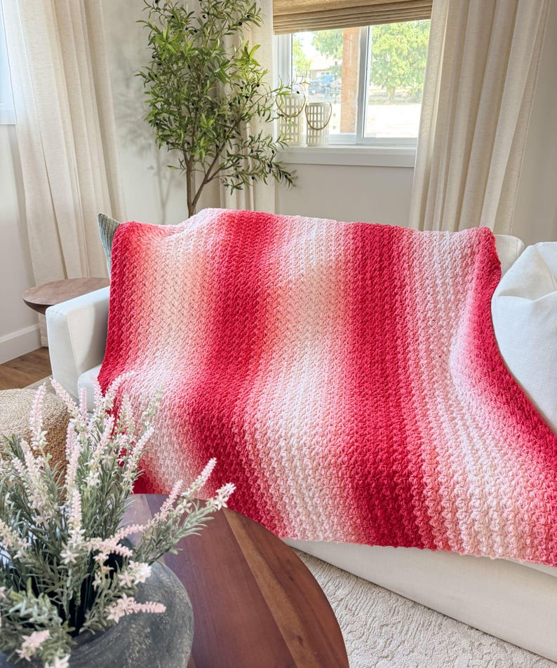 May include: A handmade, textured blanket in shades of red and pink, draped over a white sofa. The blanket features a gradient effect, transitioning from light pink to a vibrant red. The cozy throw adds a touch of warmth and color to the living space.