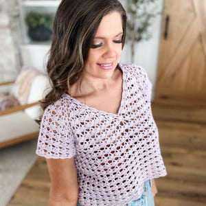 May include: A light purple, short-sleeved crochet top with a V-neck. The open weave design of the top is visible. The top is paired with blue jeans.