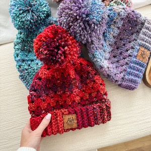 May include: Three crocheted hats with pom-poms. The hats are in shades of blue, purple, and red. The red hat has a brown leather tag with the text "Mel's Hand-Crochet Designs".