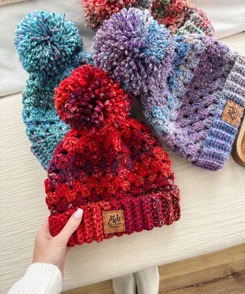 May include: Three crocheted hats with pom-poms. The hats are in shades of blue, purple, and red. The red hat has a brown leather tag with the text "Mel's Hand-Crochet Designs".