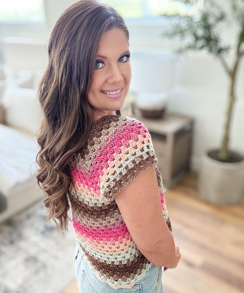 May include: A short-sleeved, crochet top in a granny square pattern. The top features horizontal stripes of brown, pink, peach, and light green. The garment is worn with blue jeans. The background is a well-lit room with a wooden floor.