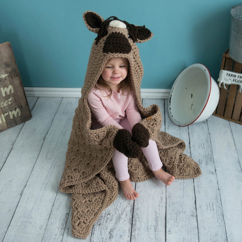 Pu&ograve; includere: Una coperta con cappuccio in crochet marrone con un design a testa di cavallo. La coperta ha dettagli in crochet marrone e bianco e un paio di guanti in crochet marrone attaccati alla coperta.