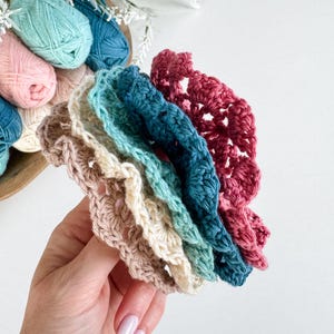 May include: A hand holds a stack of colorful, crocheted coasters in shades of pink, teal, blue, and cream. The coasters have a scalloped edge and are made of yarn. A wooden bowl with yarn balls in various colors is in the background.