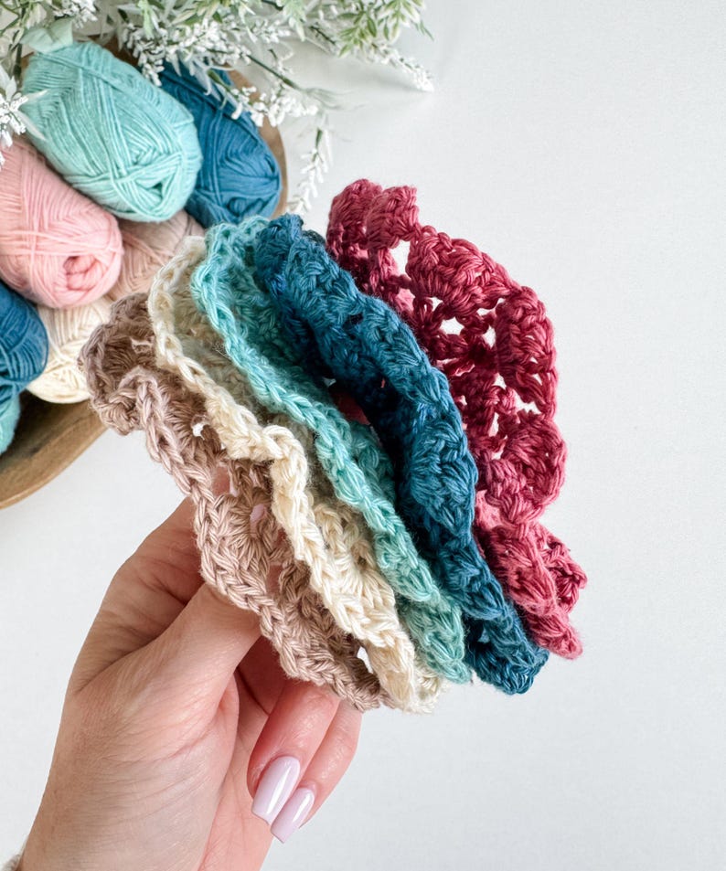 May include: A hand holds a stack of colorful, crocheted coasters in shades of pink, teal, blue, and cream. The coasters have a scalloped edge and are made of yarn. A wooden bowl with yarn balls in various colors is in the background.