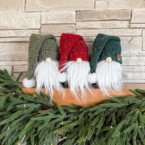 May include: Three gnomes with white beards and knitted hats. The hats are green, red, and green. The gnomes are sitting on a wooden surface with a green garland in front of them.